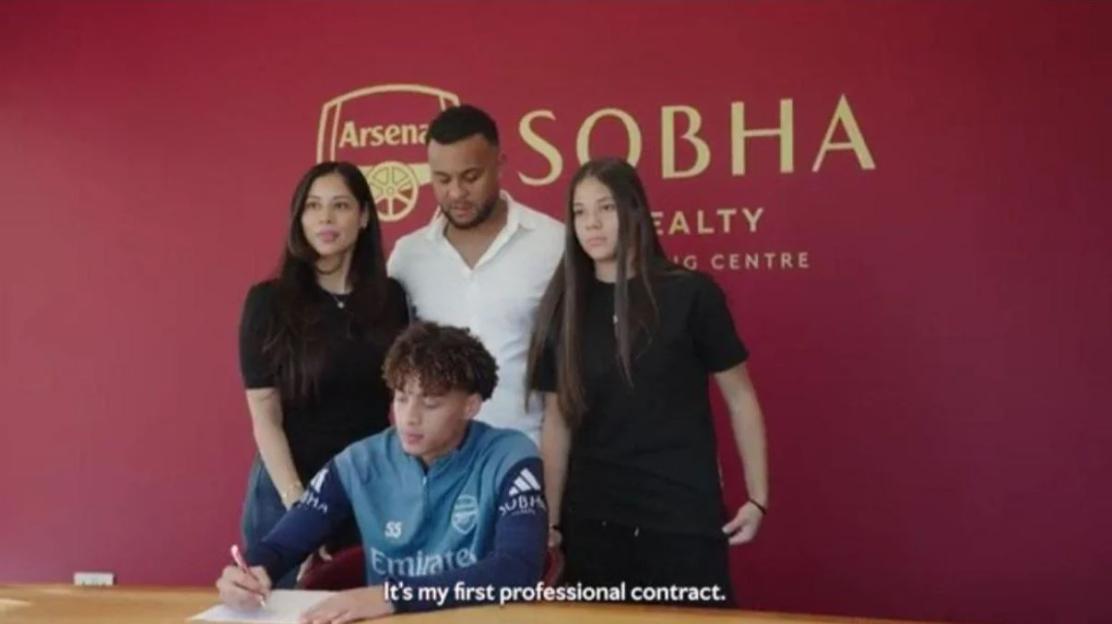 A young man signing his first professional contract, surrounded by his family and an Arsenal logo.