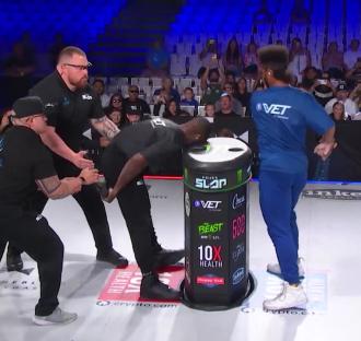 A man bending over a cylindrical object, being held up by two security personnel, while another person stands nearby.