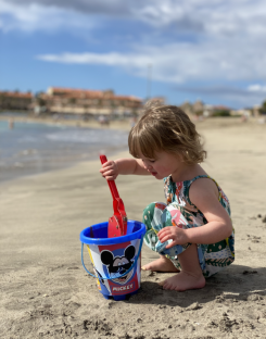 Toddler playing with a Mickey Mouse bucket and sand shovel on a beach.