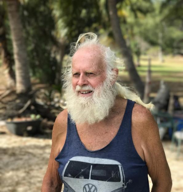 Smiling older man with long white hair and beard wearing a blue tank top.
