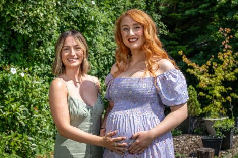 A pregnant woman and her friend stand outside, smiling.