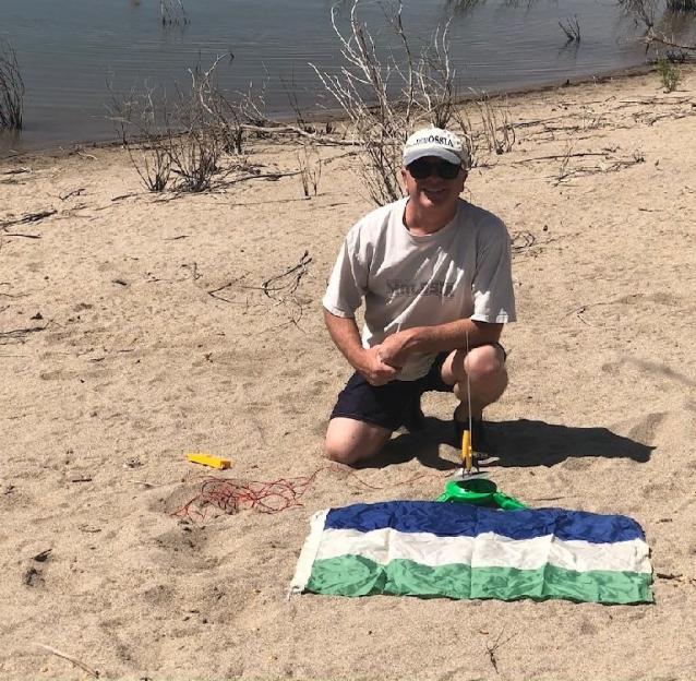 A man kneels on a beach near a lake, preparing to raise the flag of Molossia, a micronation. A person kayaks in the background.