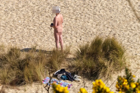 A nude person standing on a sandy beach with clothing nearby.