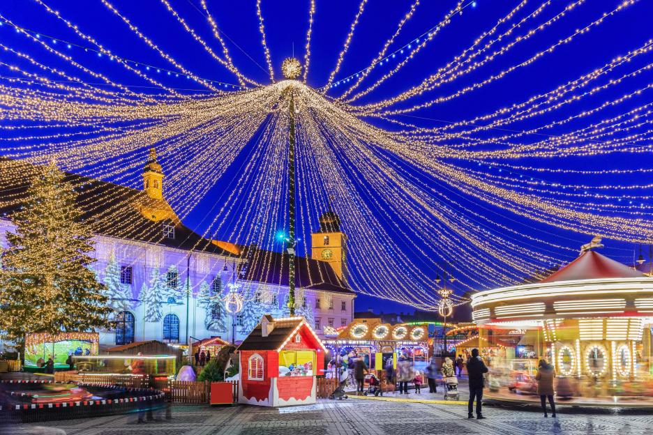 Christmas Market at twilight in Sibiu, Romania, with people walking around a carousel and small shops under strung lights.