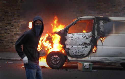 A hooded youth stands near a burning van during a riot.