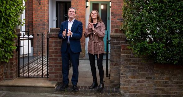A man and a woman standing outside a house, clapping.