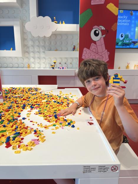 A boy holding a yellow and blue Lego creation while sitting at a table covered in Lego bricks.
