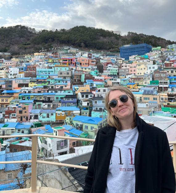 A woman with sunglasses stands in front of a hillside village of colorful houses.