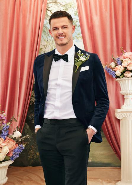 Steven from Married At First Sight, wearing a dark blue suit jacket with a black bow tie and white shirt, stands smiling in front of a draped coral background with floral arrangements.