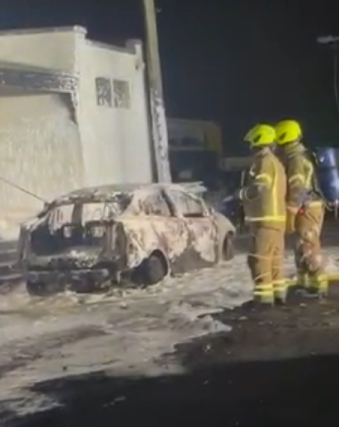 Two firefighters stand next to a burnt-out car covered in white fire retardant foam at night.