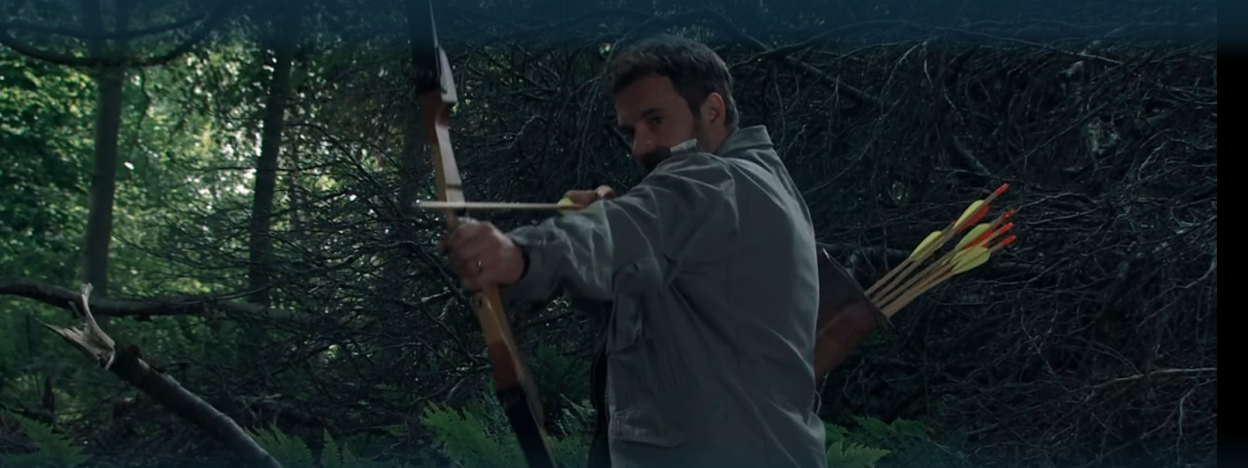 Man aiming a bow and arrow in a forest.
