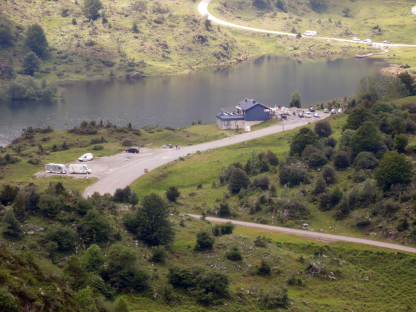 Aerial view of a lake, building, and parked vehicles in a mountainous area.