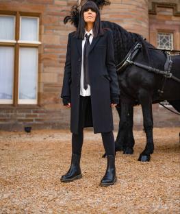 Claudia Winkleman in a black coat standing by a black horse.