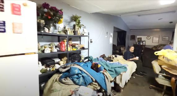 Interior of a cluttered home with a man sitting on a bed.