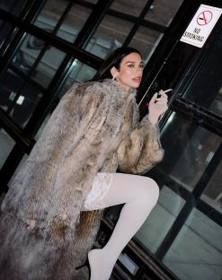 Dua Lipa in a fur coat and white stockings, holding a cigarette.