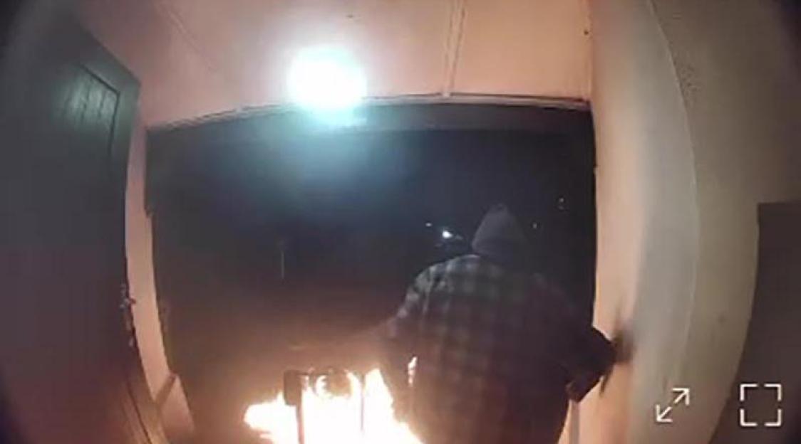 A figure in a plaid shirt and dark hoodie stands near a building entrance with a fire on the ground.