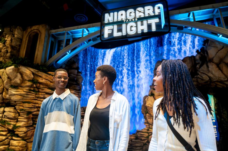 Three people stand before a Niagara Falls Takes Flight sign and simulated waterfall.
