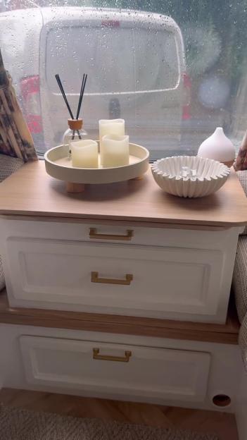 Close-up of a caravan interior with decorative items, including a reed diffuser, three flameless candles on a tray, and a white rippled bowl, on a wooden countertop above white drawers with gold handles.