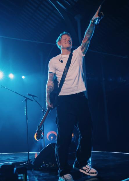 Ed Sheeran performing live with a guitar on stage, bathed in blue light, pointing his arm up with a smile, in front of a "Billions Club Live" sign.