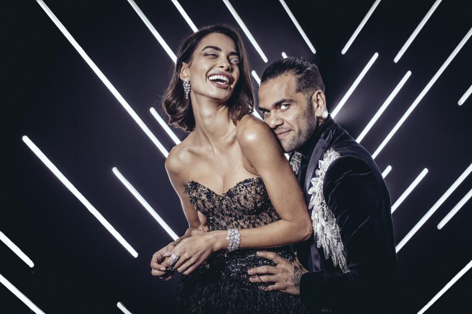 Dani Alves and Joana Sanz posing in a photo booth with a black background and diagonal white lights.