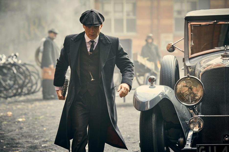 Cillian Murphy as Tommy Shelby in a long black coat and flat cap, walking past a vintage car.