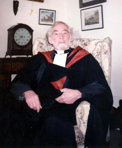 The Reverend Ronald Glazebrook sitting in a patterned armchair, wearing glasses, a white clerical collar, and a black robe with a red stole.