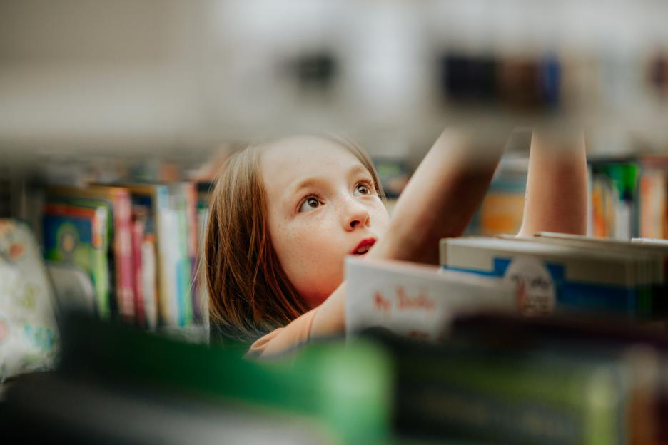 A young girl with freckles and brown hair reaching for a book on a library shelf.