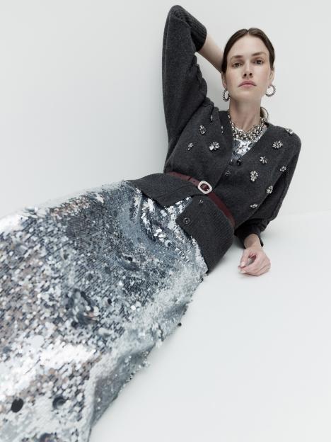 A model wears a gray cardigan with jewel accents, a silver sequin top, a silver sequin skirt, and a thin brown belt.