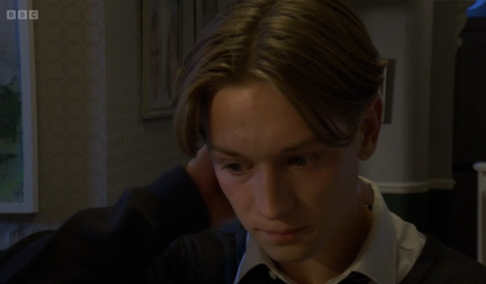 A young man with light brown hair looking down, with his hand touching his neck.