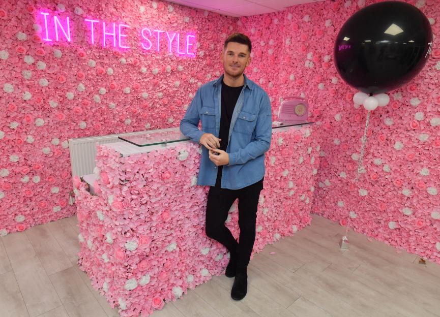 Adam Frisby, founder of 'In The Style,' standing in front of a pink floral wall with a neon sign that reads "IN THE STYLE."