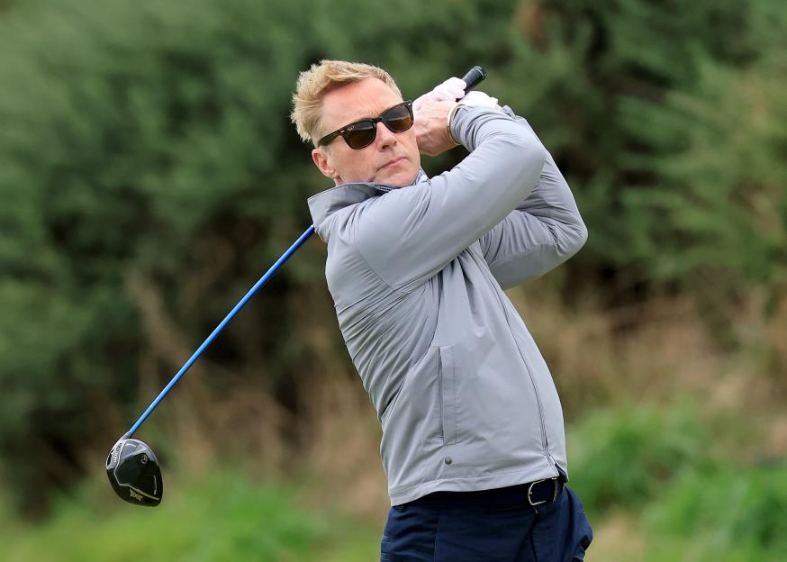 Ronan Keating wearing sunglasses and a grey jacket, mid-swing with a golf club.