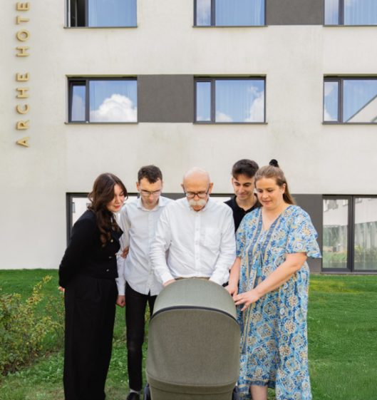 Five people gathered around a baby carriage on a lawn outside a building.