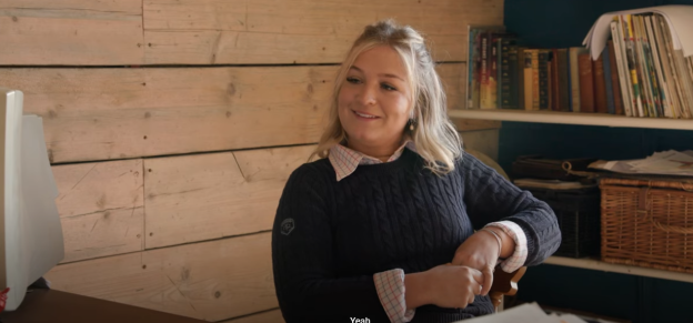 A woman sits at a desk in a wooden-paneled room, smiling.