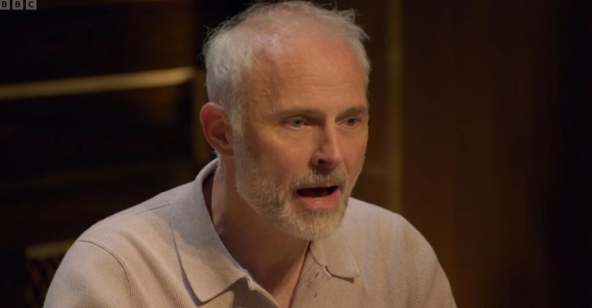 Close-up of a middle-aged man with grey hair and beard, wearing a light-colored collared shirt, looking slightly to the right with his mouth open.