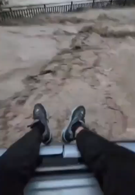 A first-person perspective of a person's legs and feet in black pants and sneakers hanging over the edge of a moving ride, with blurred sandy terrain and a fence in the background.