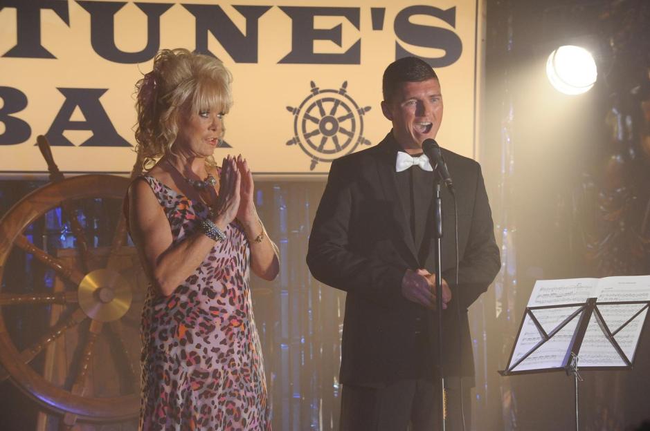 Asa Elliott singing into a microphone on stage, with a woman in a leopard-print dress next to him, and a sign behind them that says "FORTUNE'S BAR" with a ship's wheel logo.