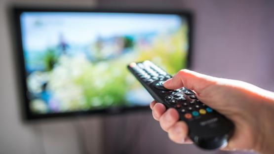 Hand holding a TV remote control in front of a blurry television screen.