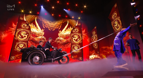 A stage performance featuring a man on a sidecar motorcycle and a man walking a tightrope.