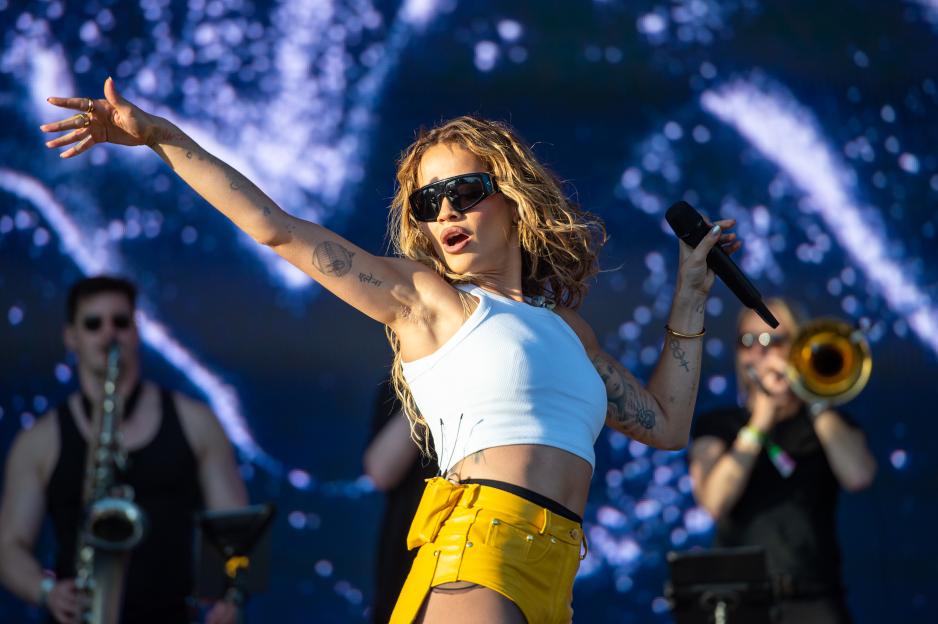 Rita Ora performs on stage in a white top, yellow shorts, and sunglasses, holding a microphone.
