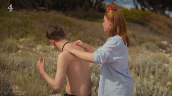 A woman applies sunscreen to a man's back on a beach.