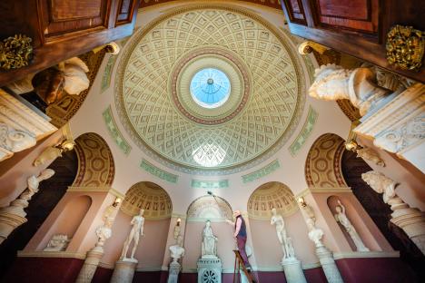 A man cleans a statue gallery with ornate ceilings and numerous classical sculptures.