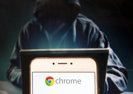 Photo illustration of a smartphone displaying the Chrome logo, with a blurred image of a hooded figure using a laptop in the background.