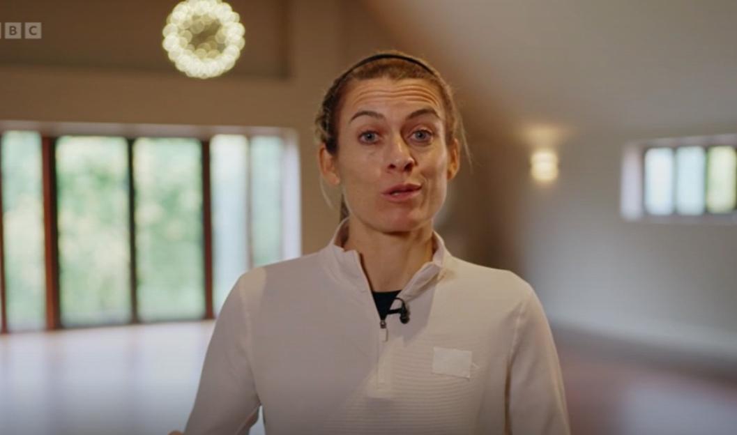 A woman in a white long-sleeved shirt with a black undershirt and a black headband speaks to the camera in a modern room with large windows.