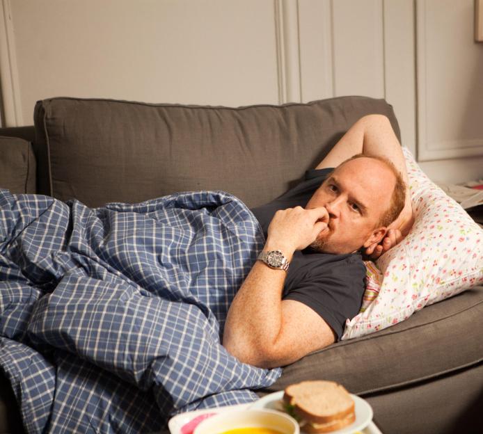 Louis C.K. lying on a sofa with a sandwich and soup in front of him.