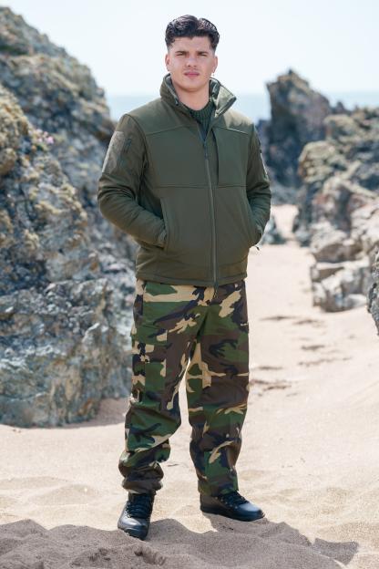Man in camouflage pants and green jacket standing on a beach.