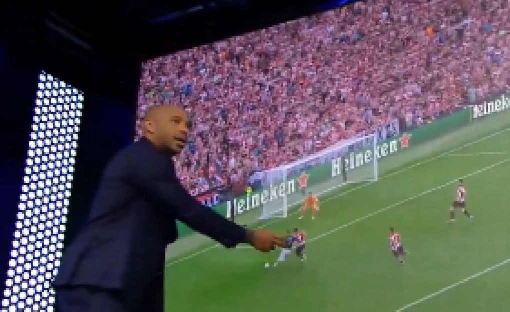 Thierry Henry pointing at a large screen displaying a football match.