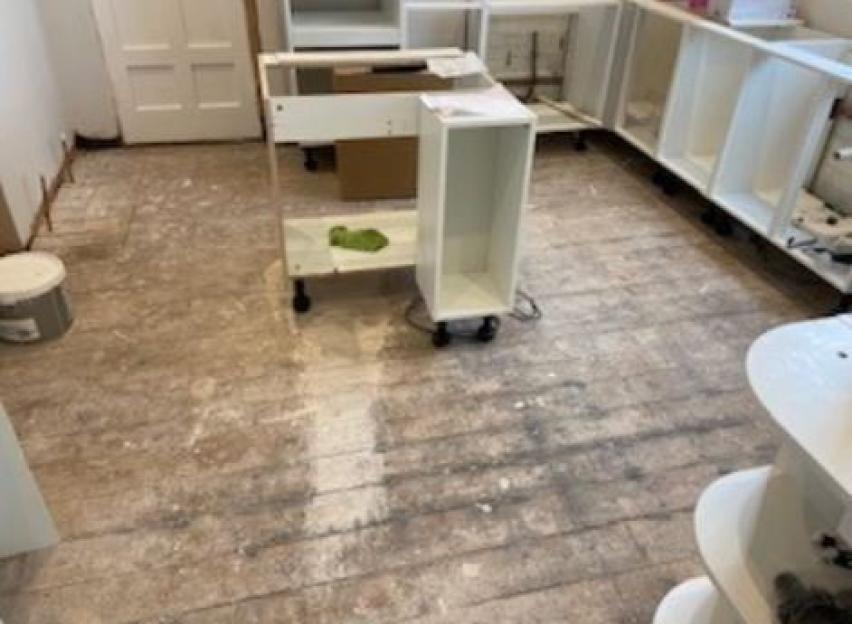 A messy construction site with exposed wooden floors, partially installed white cabinets, and building materials.