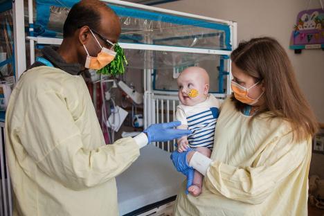 Photo of a baby in a hospital room with two medical professionals.