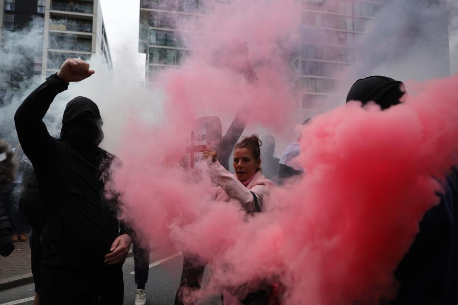 Anti-migrant protestors clash with police amidst pink smoke bombs.