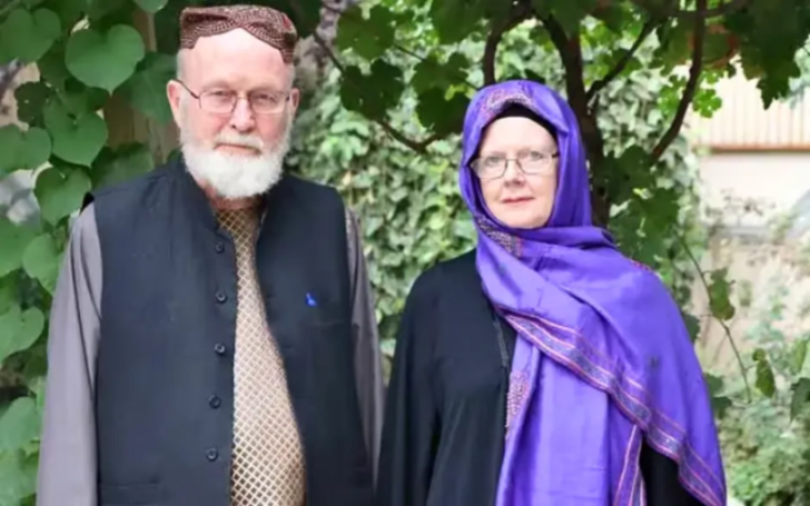 A man in a skullcap and black vest with a beard and glasses next to a woman in a purple hijab and glasses.